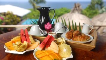Un desayuno con diversas frutas y pasteles sobre una bandeja de madera, una jarra y tazas en una mesa con vistas panorámicas al fondo.