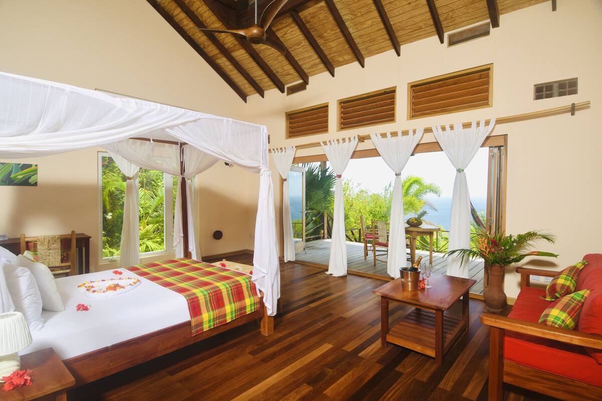 A spacious room with a bed, a wooden table, and a red sofa. There are windows with curtains, a ceiling fan, and a view of the outdoors.