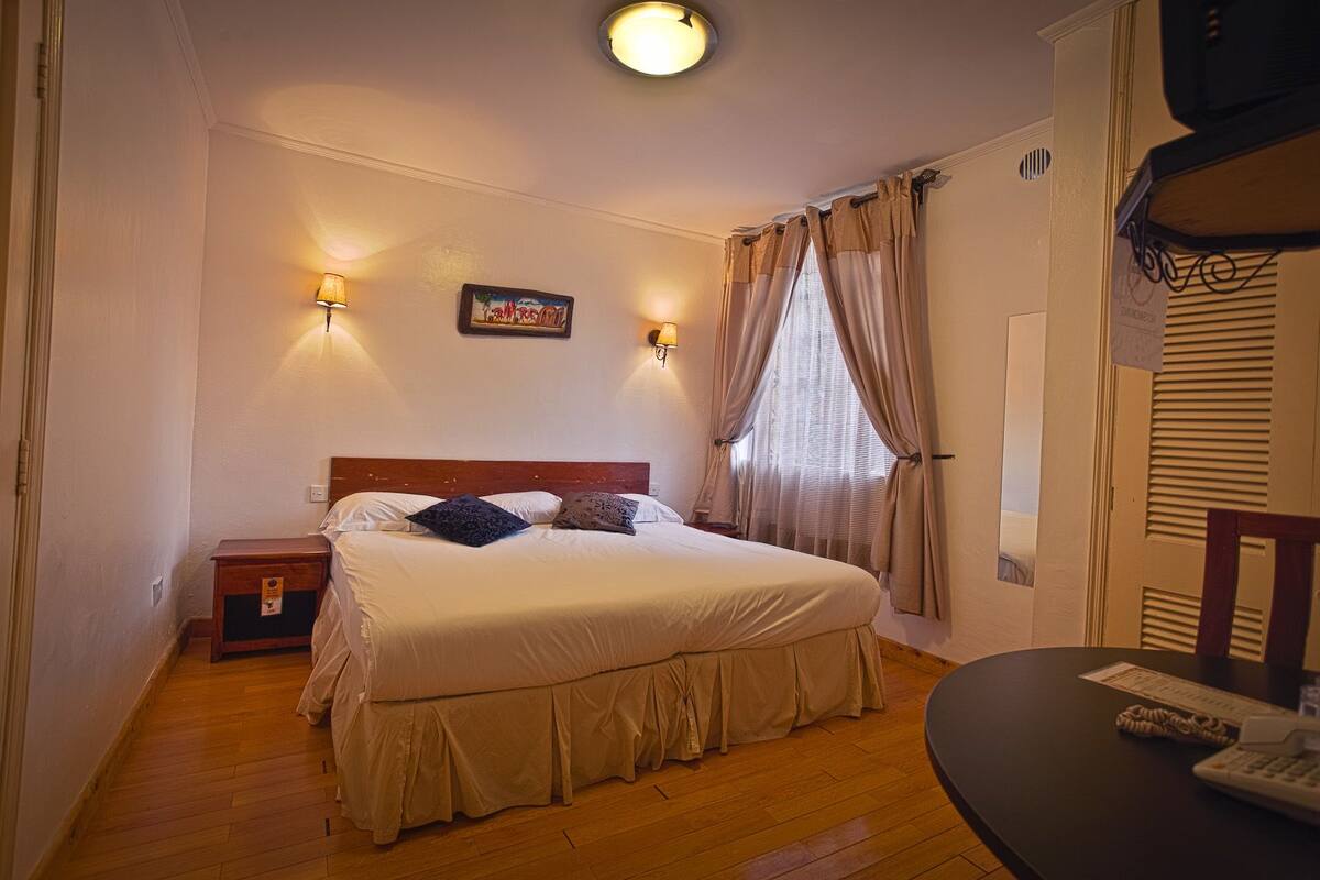 Un dormitorio ordenado con una cama, lámparas de noche, una mesita y una ventana con cortinas.