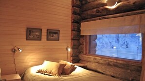 Une chambre confortable dans un chalet en rondins, avec un lit, deux lampes fixées au mur et une fenêtre avec des stores.
