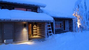 Une cabane en rondins dont le toit est recouvert de neige, avec une échelle en bois menant à l’entrée.