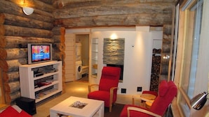 L'intérieur d'une cabane en rondins, avec une télévision, des étagères blanches, des chaises rouges, une table basse blanche et une cheminée.