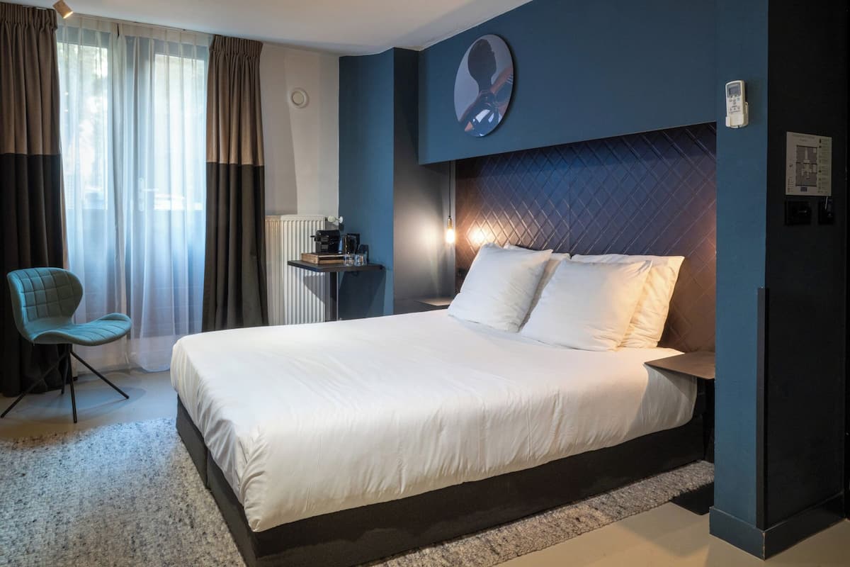 Habitación de hotel con una cama grande, una silla, una mesita de noche y una lámpara de pared.