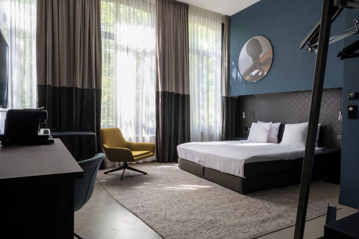 Una habitación de hotel moderna con cama, escritorio, silla y ventanas grandes con cortinas.