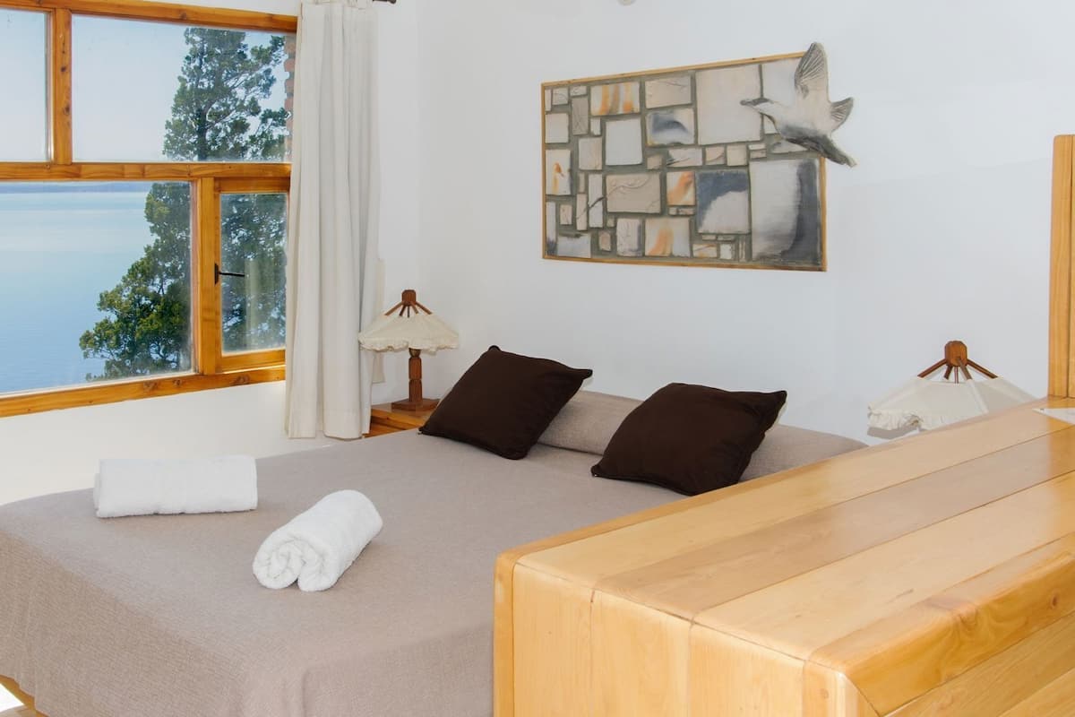 Ein Schlafzimmer mit Bett, einem Nachttisch aus Holz, einer Lampe, einem Wandbild und Blick durch ein Fenster.