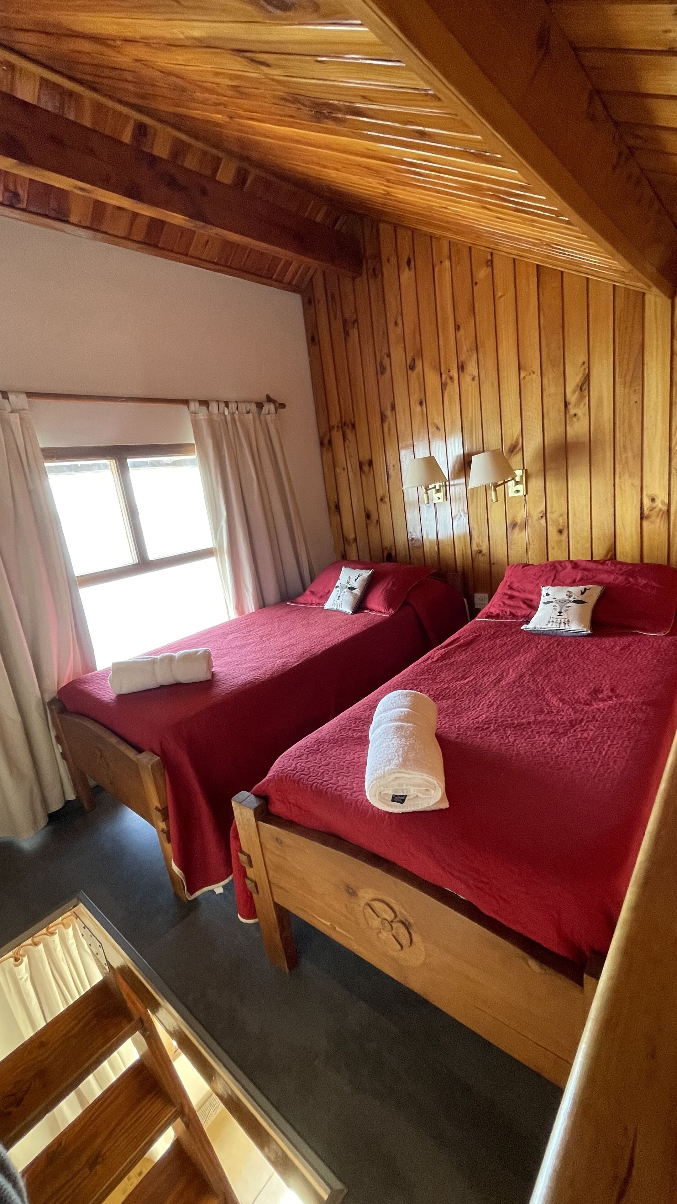 Una habitación con dos camas de madera, cada una con una colcha roja y una toalla blanca. Se observa una ventana con cortinas, techo de madera y una pequeña mesa con una silla.
