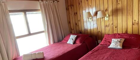 Una habitación con dos camas de madera, cada una con una colcha roja y una toalla blanca. Se observa una ventana con cortinas, techo de madera y una pequeña mesa con una silla.