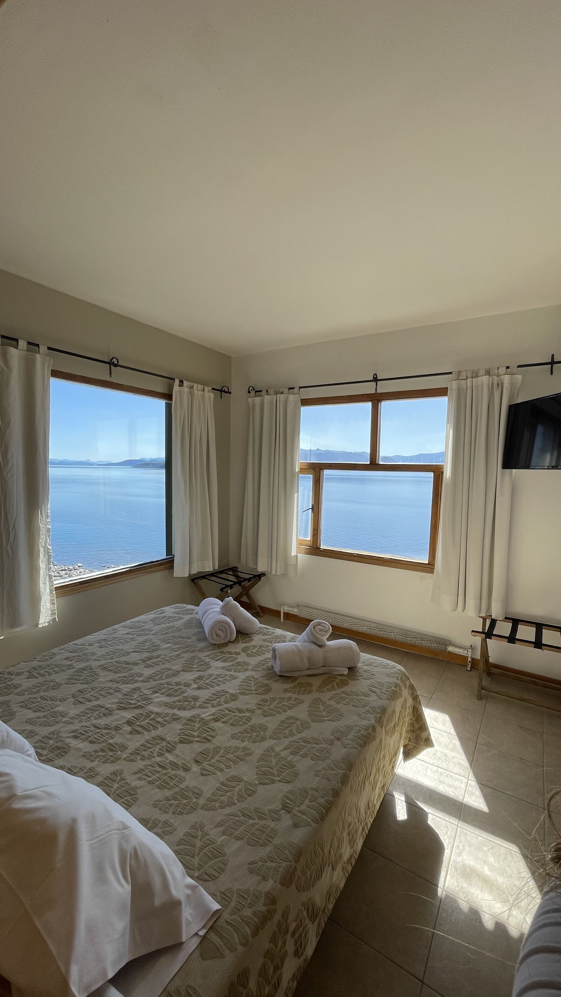 Un dormitori amb un llit gran, dues finestres amb cortines, un televisor de pantalla plana i vistes al mar.