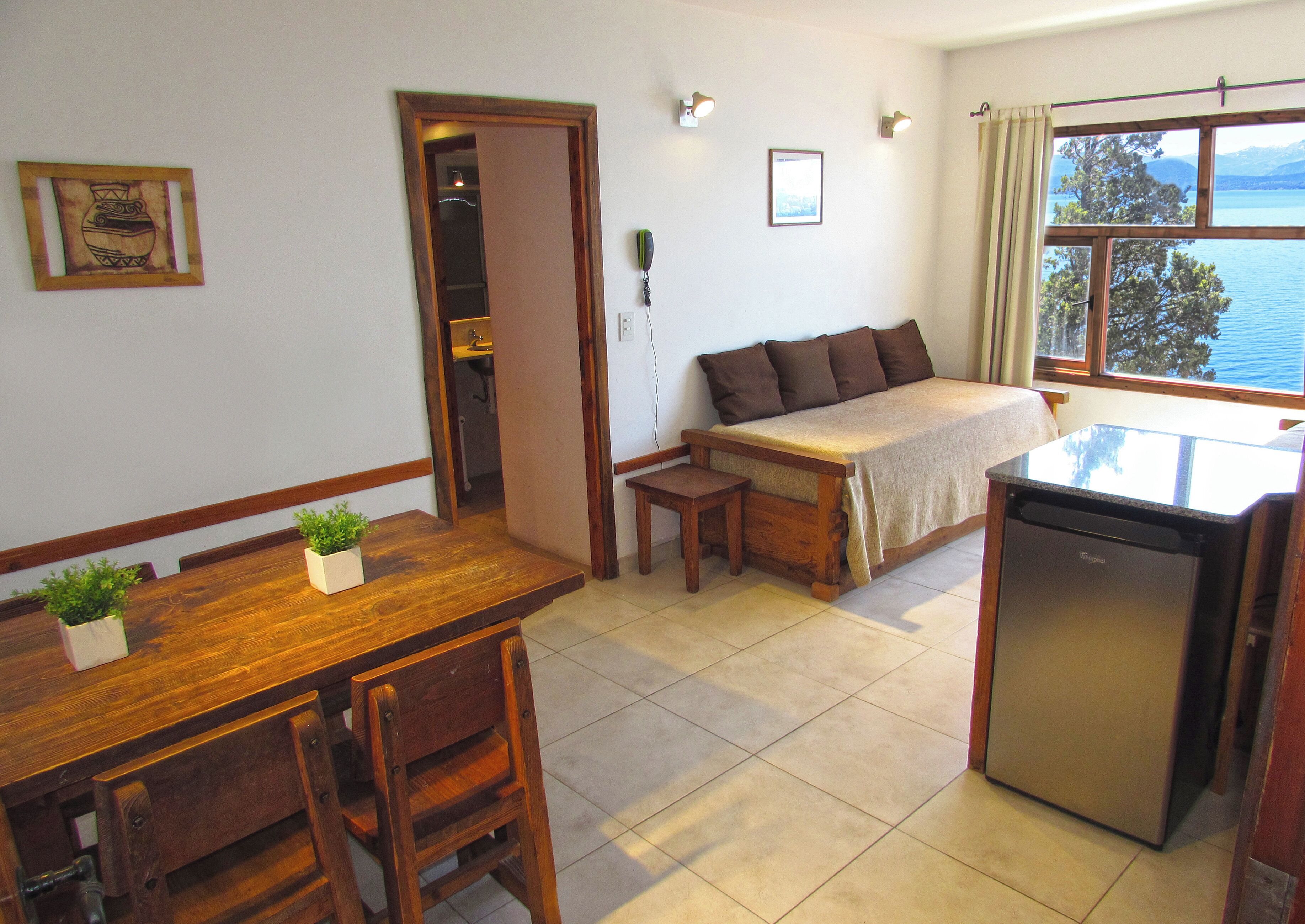 A compact room with a bed, a small table, a bench, a refrigerator, and a window with a view of the sea.
