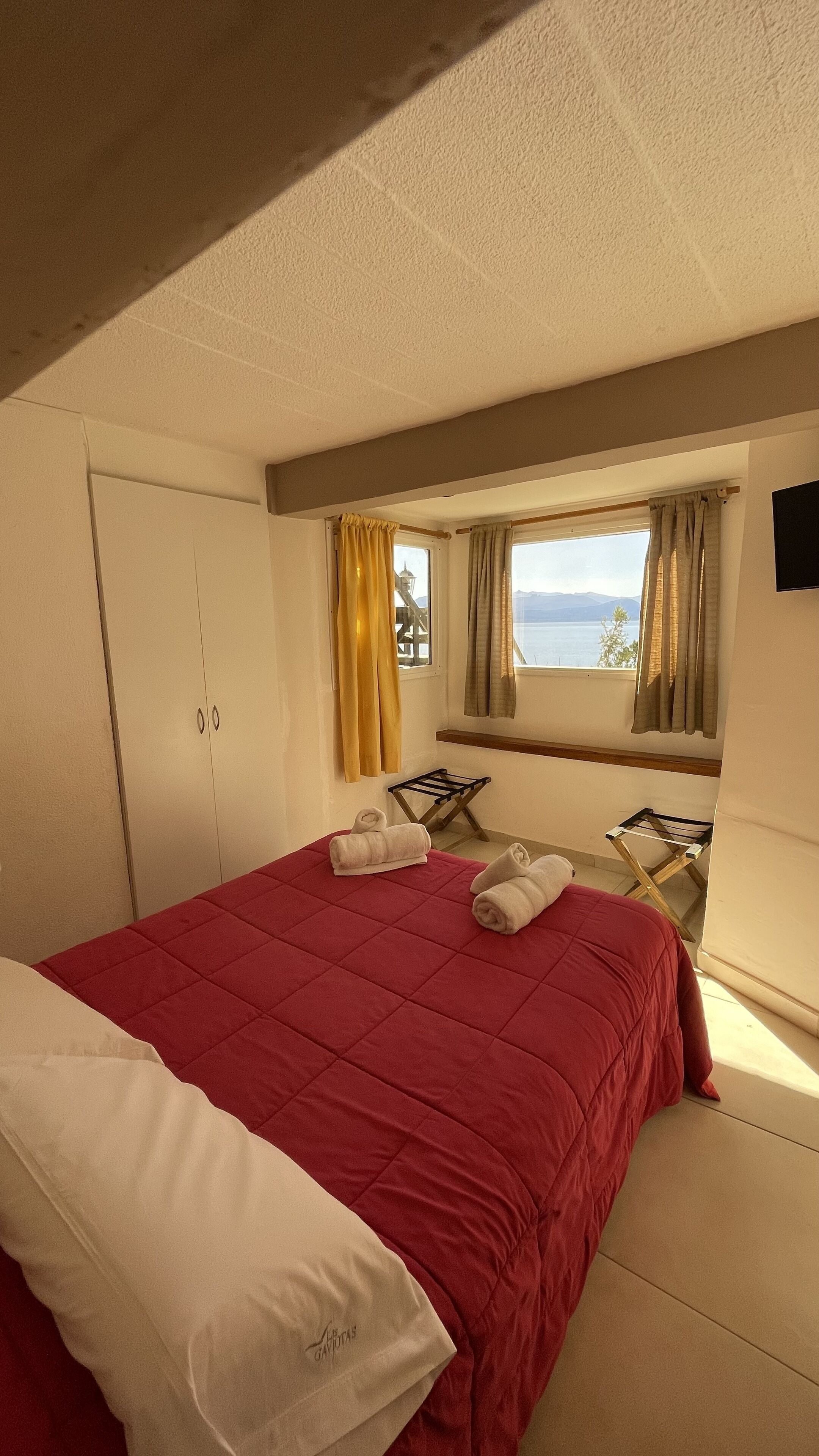 Un dormitorio con colcha roja, almohada blanca, banco de madera y ventana con cortinas.