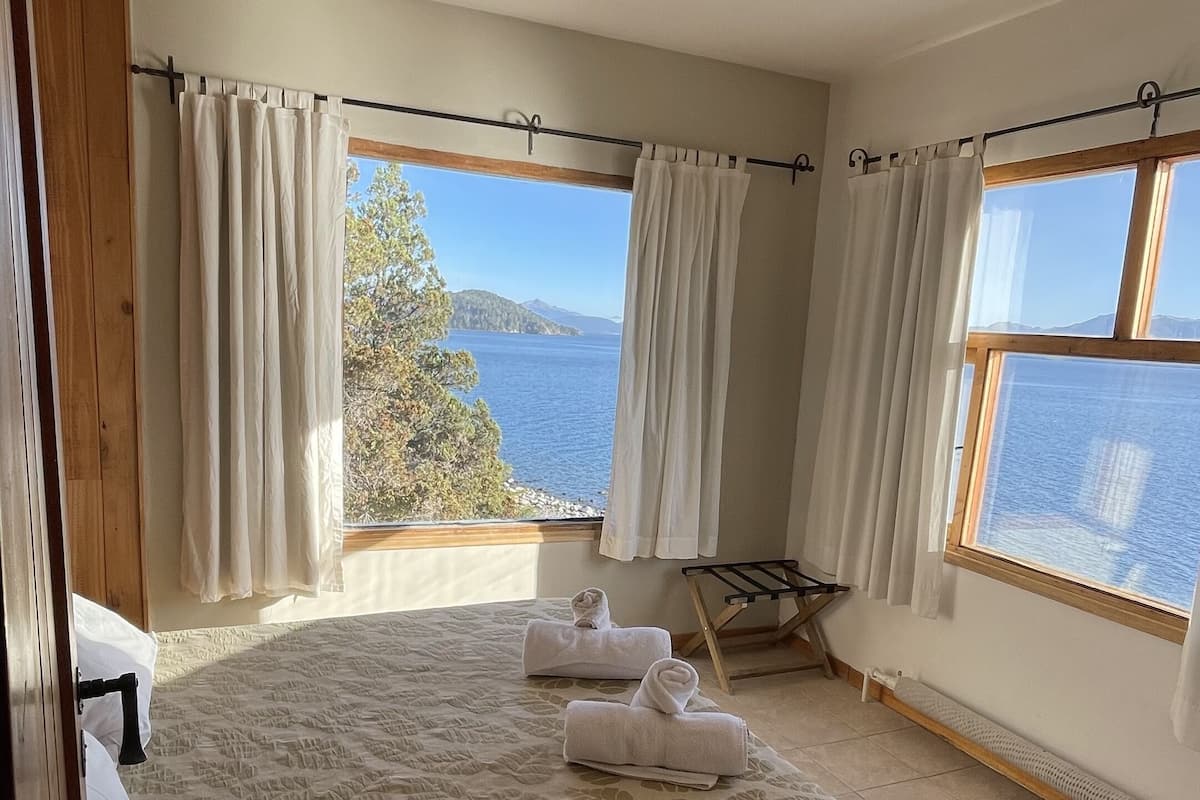 Ein Schlafzimmer mit Bett, zwei mit Vorhängen versehenen Fenstern, Blick auf das Meer und Holzboden.