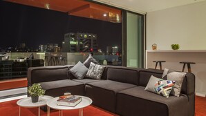 A modern living room with a large window, a grey sofa, a small round table, and a potted plant. - 1866 Court & Suites (Beirut)