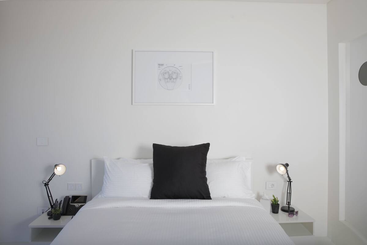 Un dormitorio minimalista con una cama, dos mesitas de noche, una obra de arte en la pared y una única almohada negra.