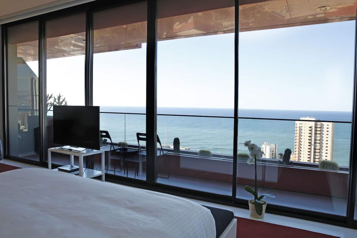 Habitación de hotel moderna con un balcón grande, una cama, un televisor y un área de comedor con vistas al mar.