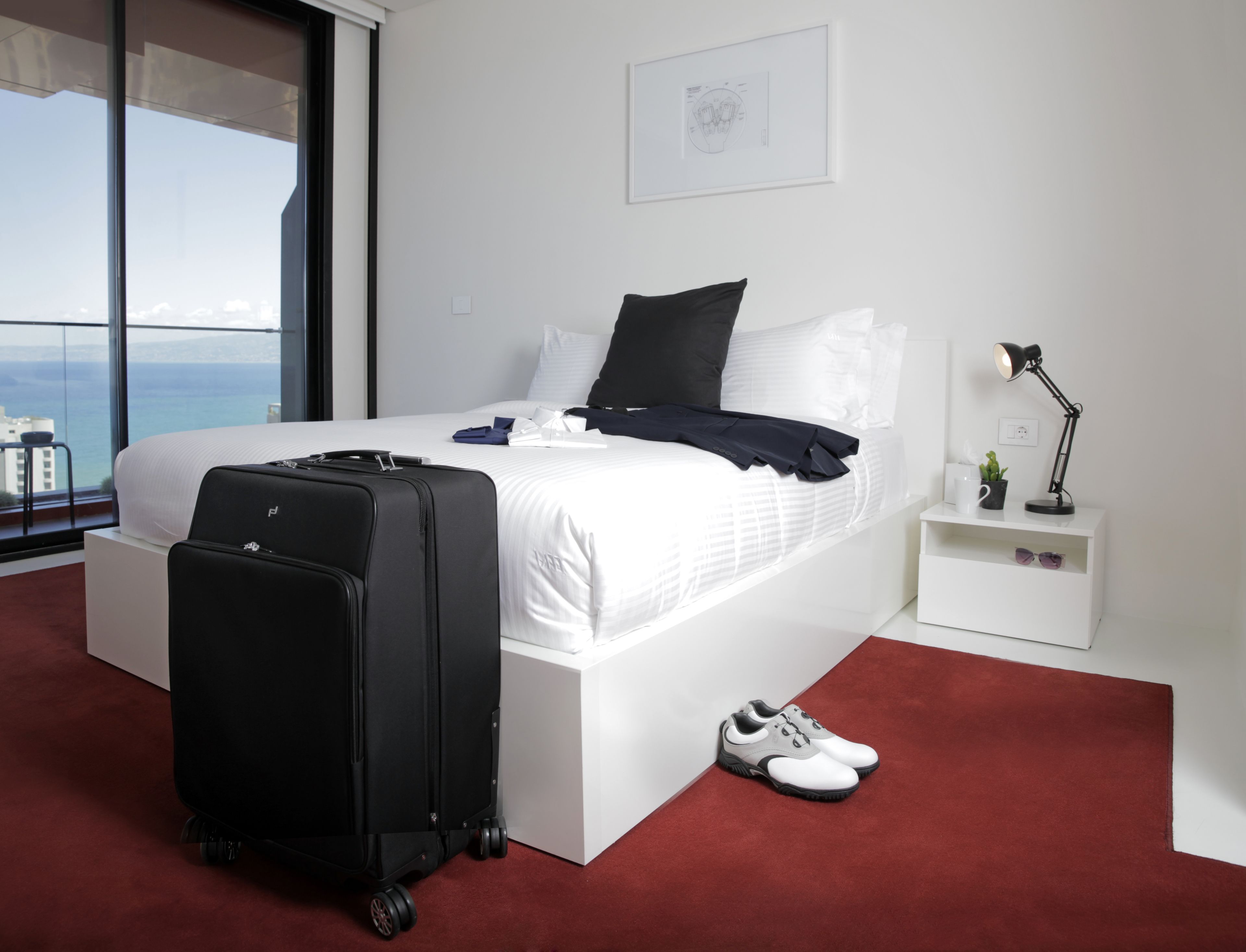 A hotel room with a large bed, a suitcase, a pair of shoes, a nightstand with a lamp, and a view of the sea through the window.