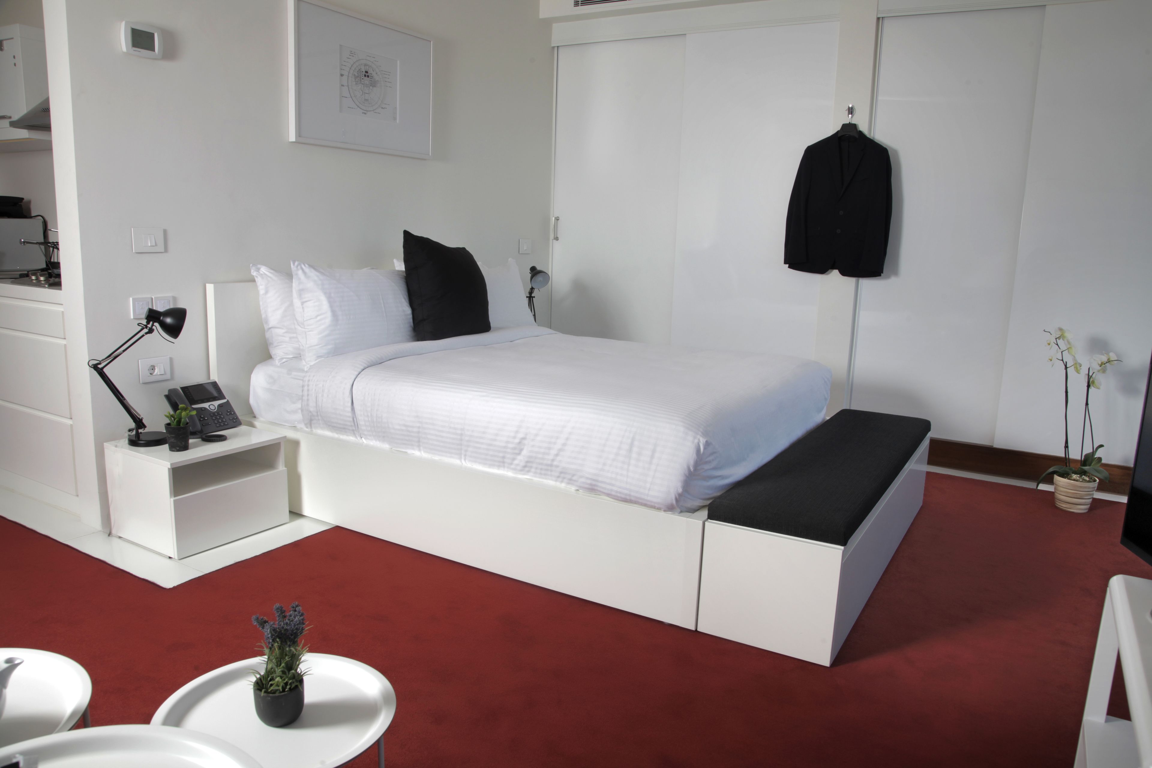 A bedroom with a white bed, a bedside table with a lamp, a small desk, a chair, and a potted plant.