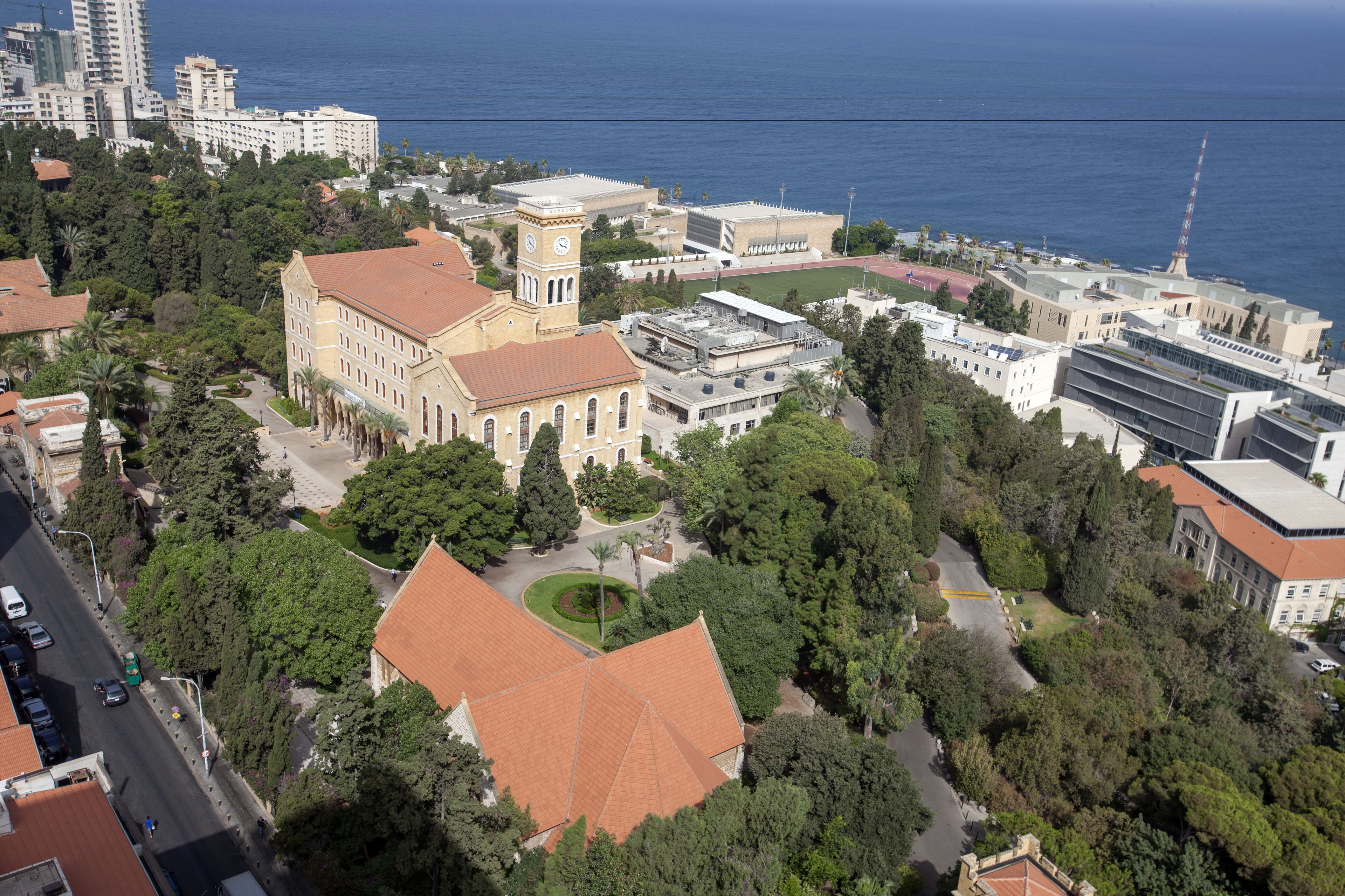 An aerial view of a campus with various buildings, including a prominent clock tower, surrounded by greenery and a body of water.