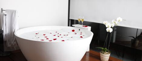 A modern bathroom with a large white bathtub filled with rose petals, a small potted orchid, and two glasses of champagne on a white side table.