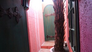 A narrow hallway with a tiled floor, a green door with a decorative metal crest, and a tree in a planter. - Afei Surf Inn - Hall 3 (Hengchun)