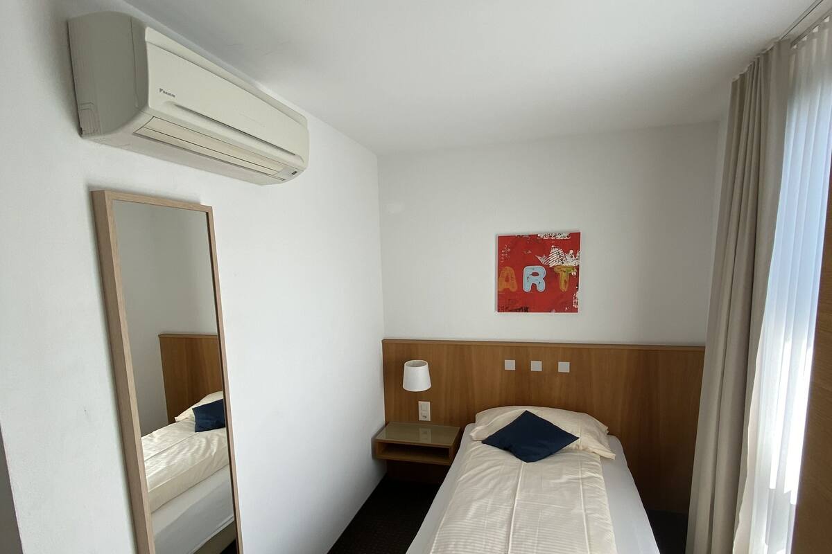 A hotel room with a single bed, a bedside table, a lamp, a mirror, an air conditioning unit, and a wall decoration.