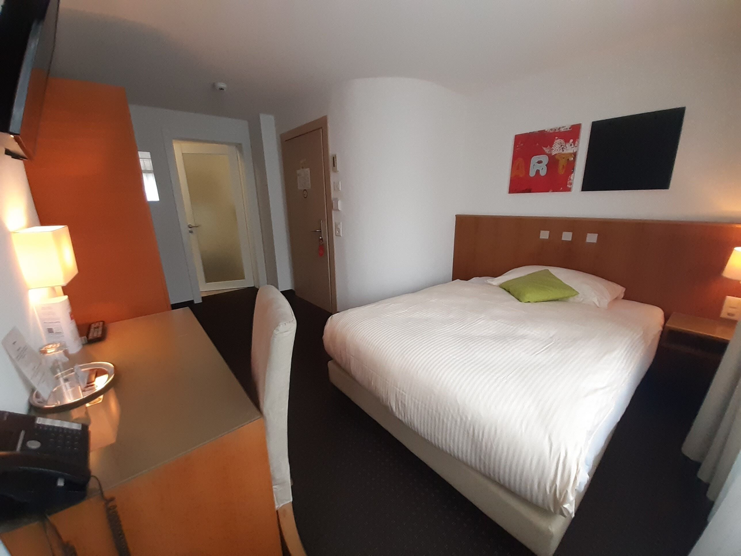 Una camera d'albergo con un letto, una scrivania, una sedia e un comodino con una lampada.