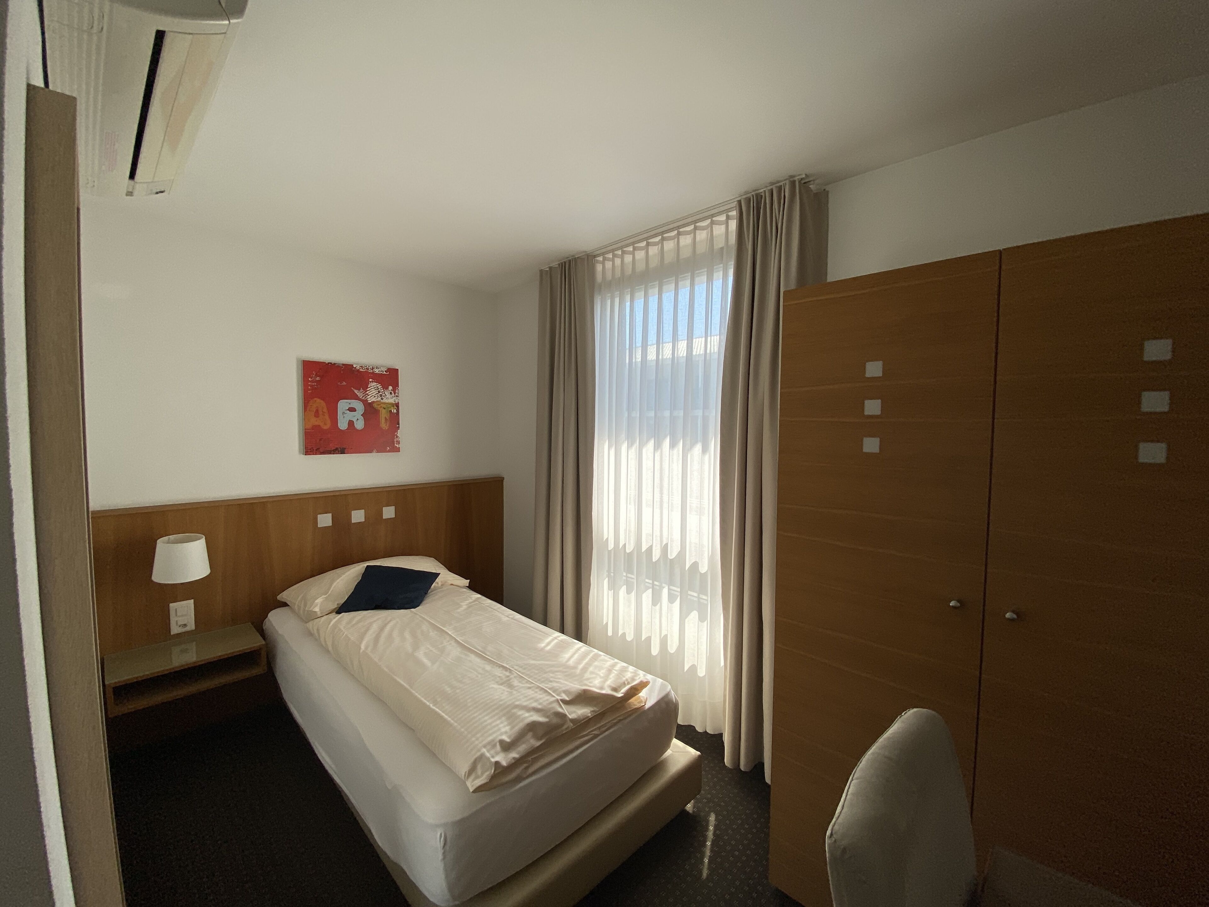A hotel room with a bed, a nightstand, a lamp, a wardrobe, and a window with curtains.