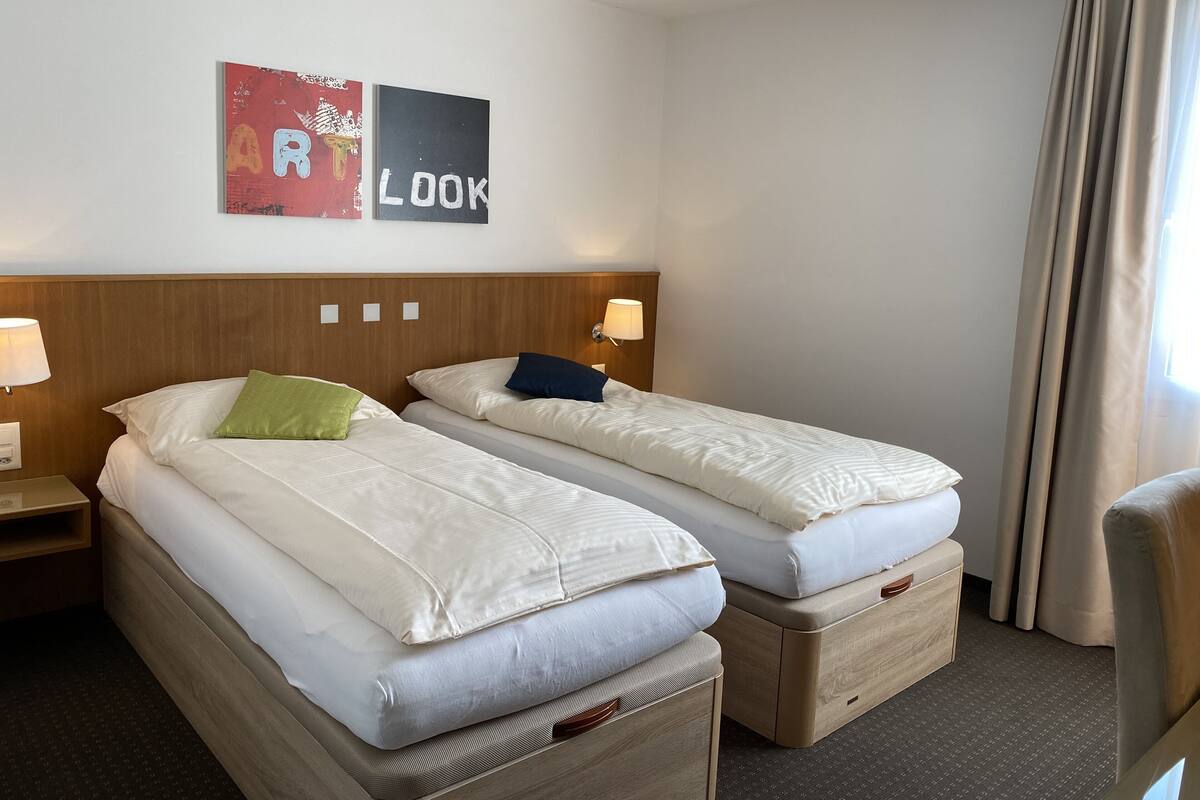 A hotel room with two beds, each with a wooden headboard, white bedding, and a green pillow on one bed. There are two bedside lamps and a wall art piece above the beds.