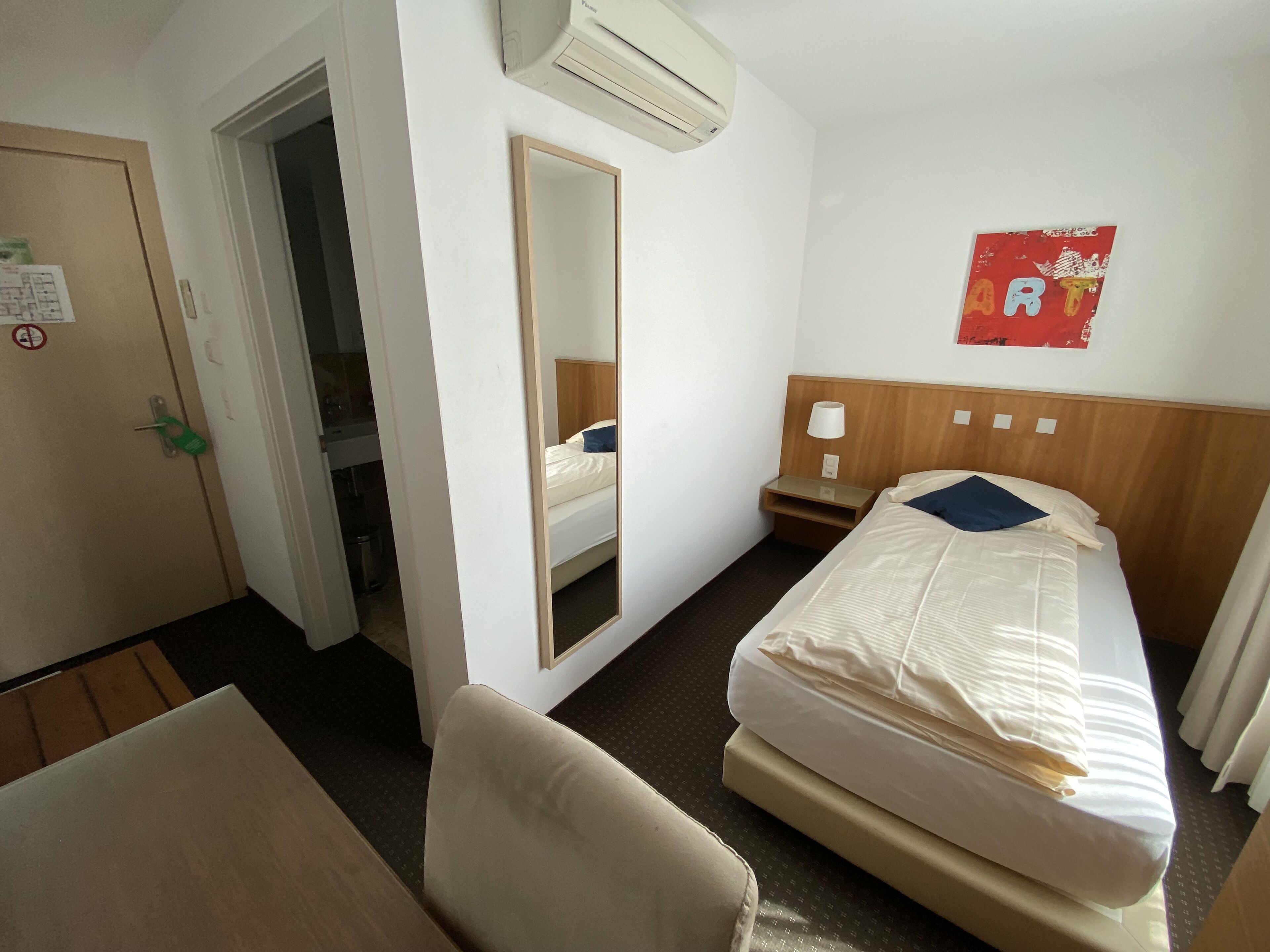 A hotel room with a bed, a chair, a desk, a mirror, and a wall-mounted air conditioner.