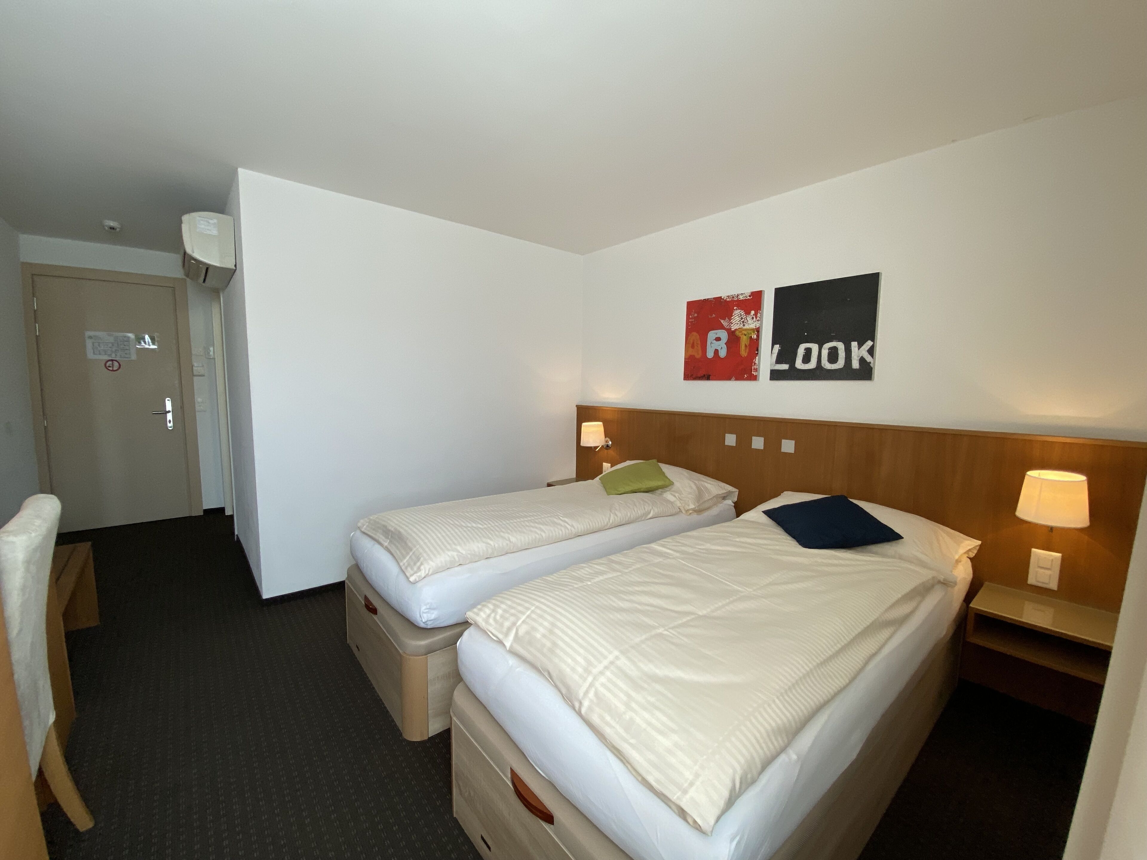 A hotel room with two beds, a desk, and a chair. There are two wall-mounted lamps and a door with a sign.