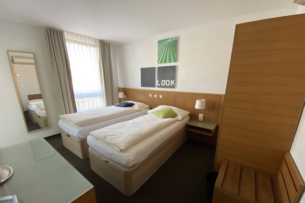 A hotel room with two single beds, a desk with a telephone, a mirror, and a wall-mounted lamp.