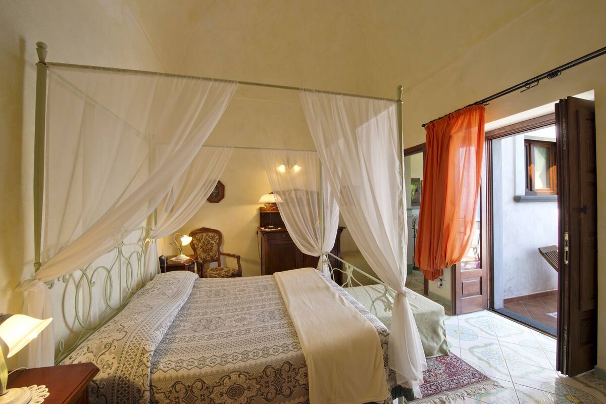 A bedroom with a four-poster bed, a nightstand, a chair, and a window with curtains.