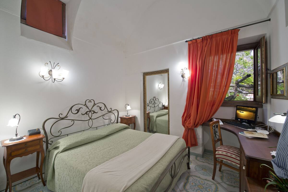 A bedroom with a bed, a desk with a computer, a chair, a nightstand with a lamp, and a window with curtains.