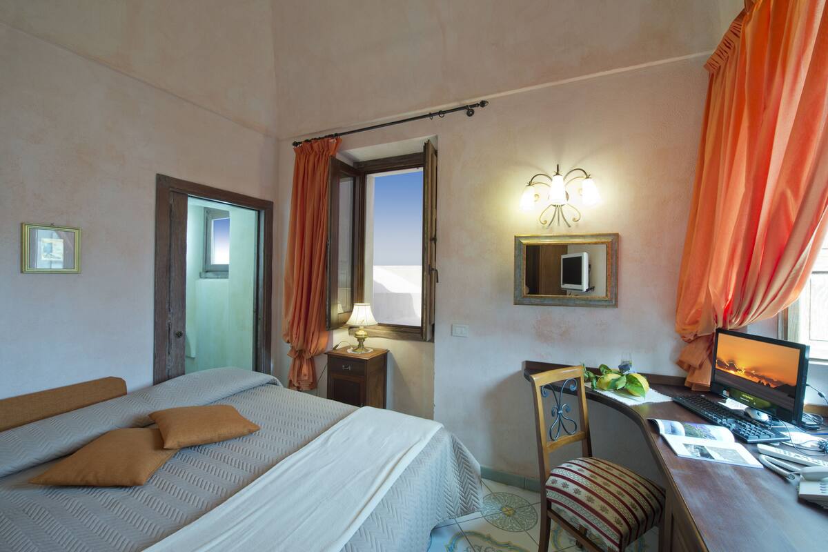 A hotel room with a bed, a desk with a computer, a chair, a lamp, and a window with curtains.