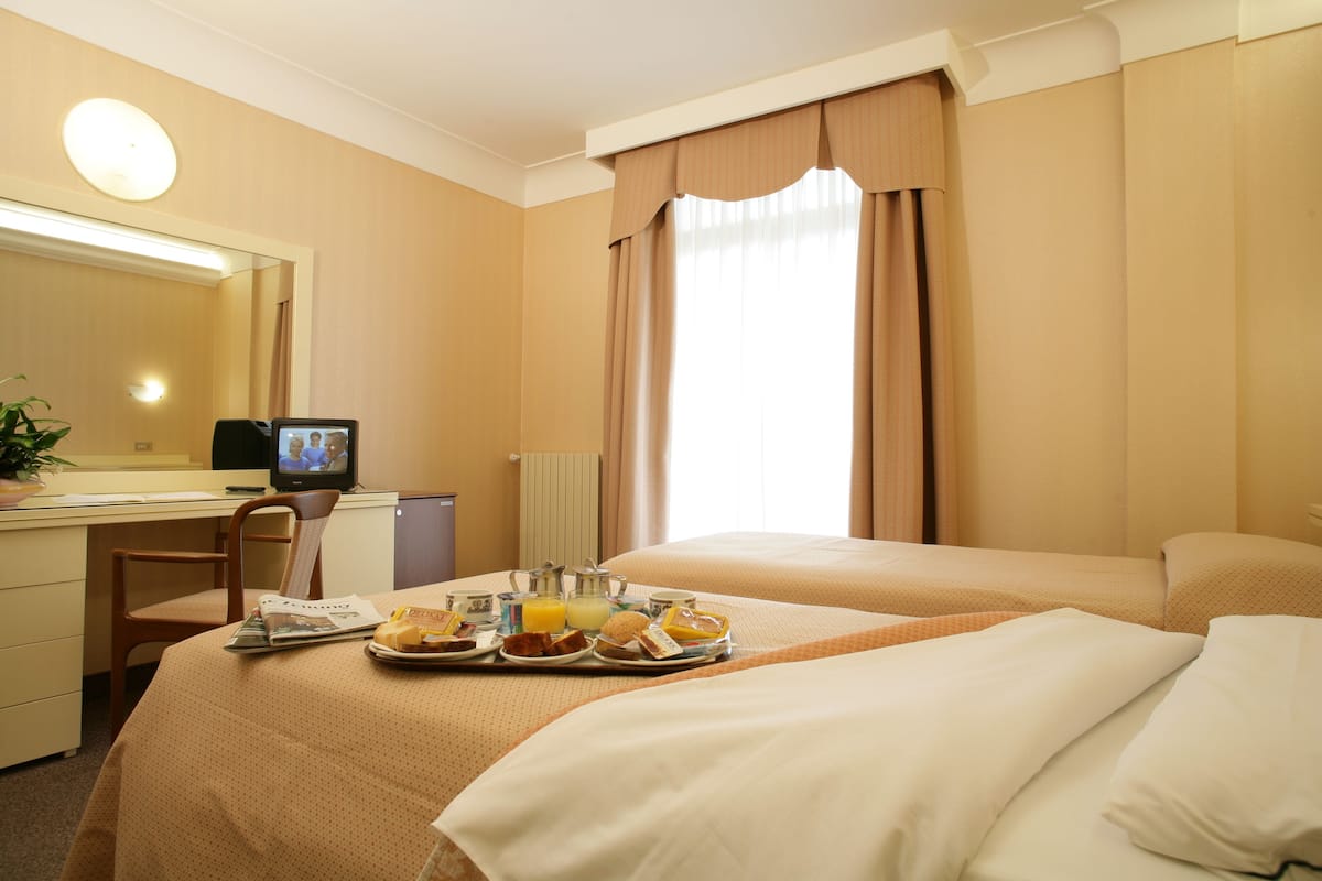 A hotel room with a bed, a desk with a computer, a tray of breakfast items, and a window with curtains.