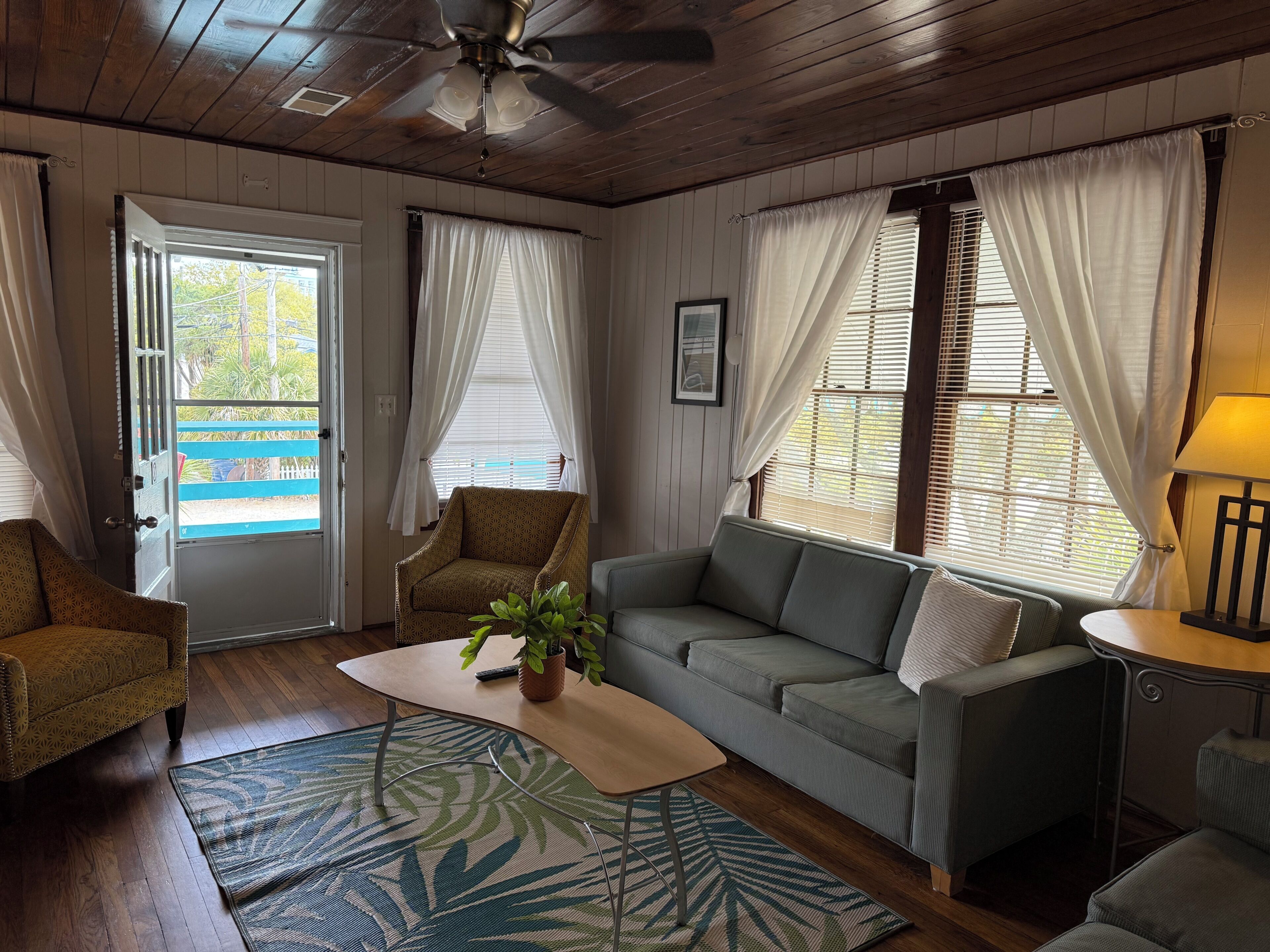 A room with a wooden ceiling, a sofa, a chair, a table with a plant, and a lamp.
