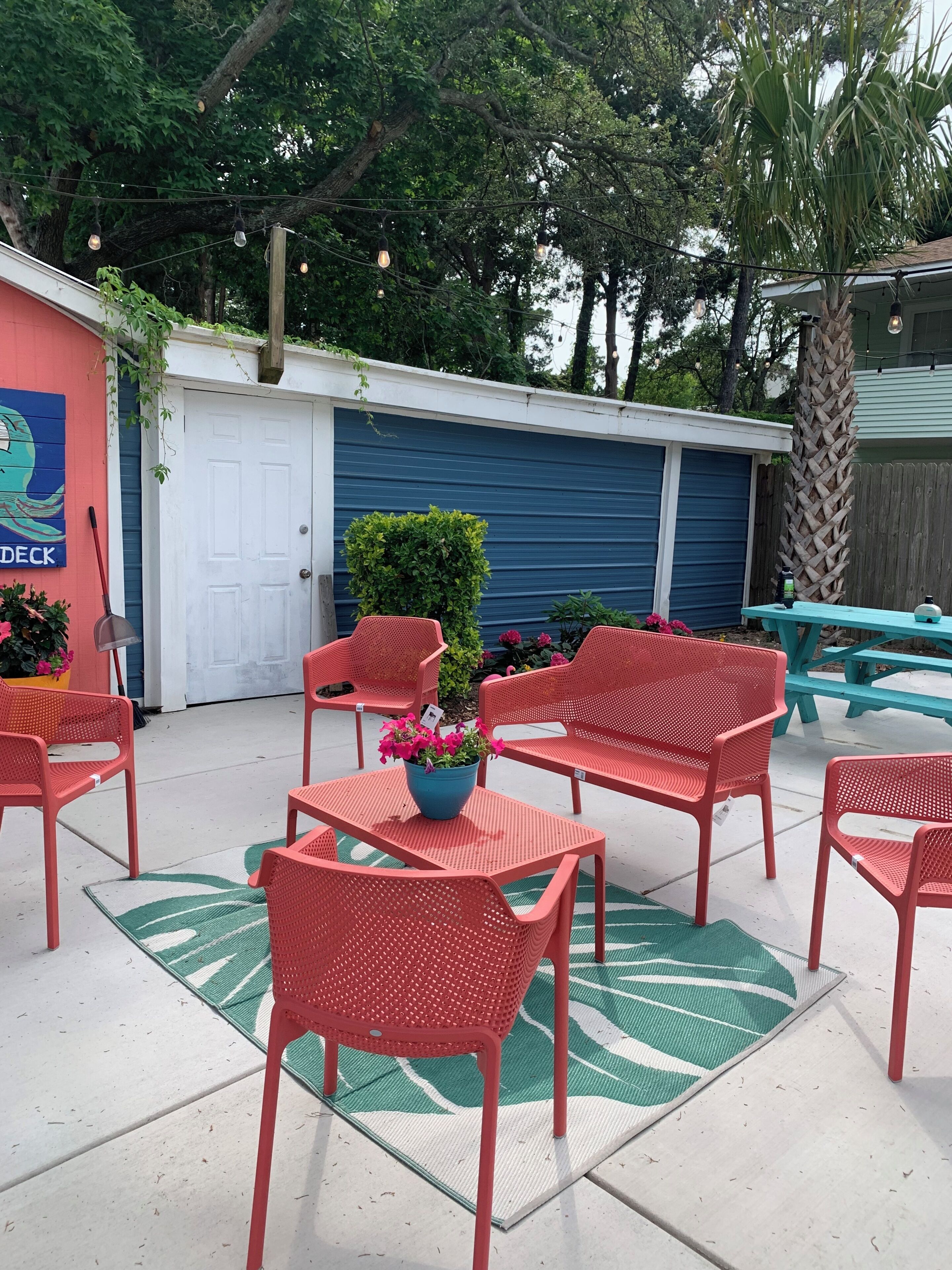 Un'area patio con sedie e tavoli in metallo rosso, una porta blu e una pianta verde come elemento decorativo.