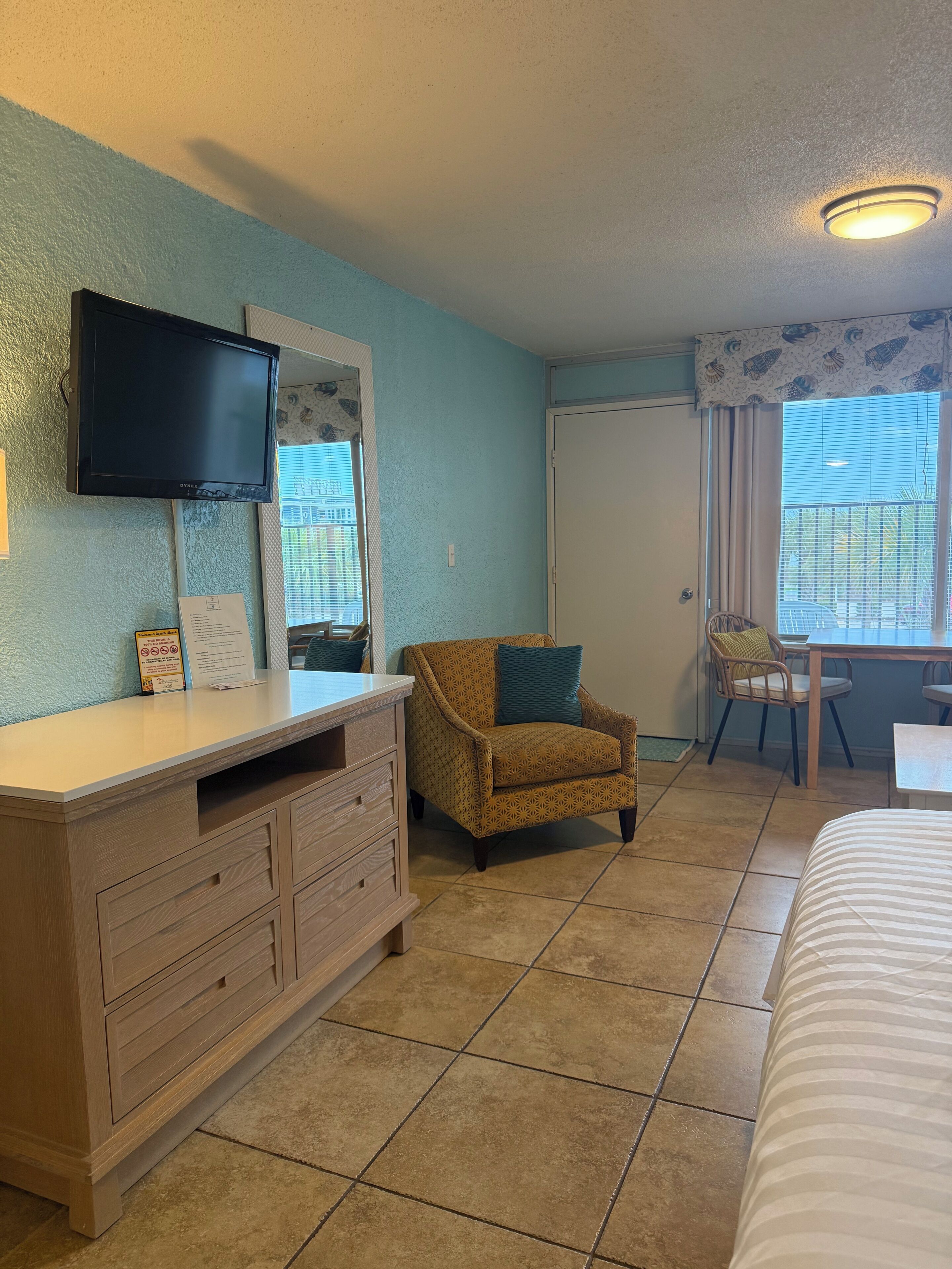 A hotel room with a bed, a dresser with a TV, a chair, a window with blinds, and a door.