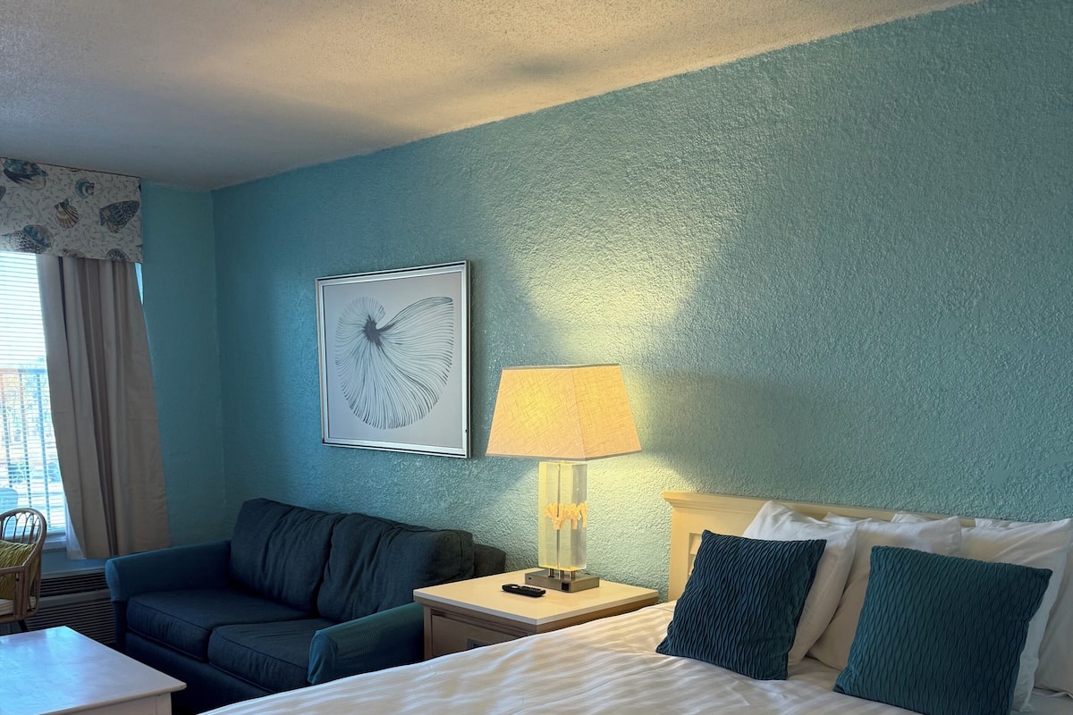 A hotel room with a bed, a sofa, a nightstand with a lamp, and a framed artwork on the wall.