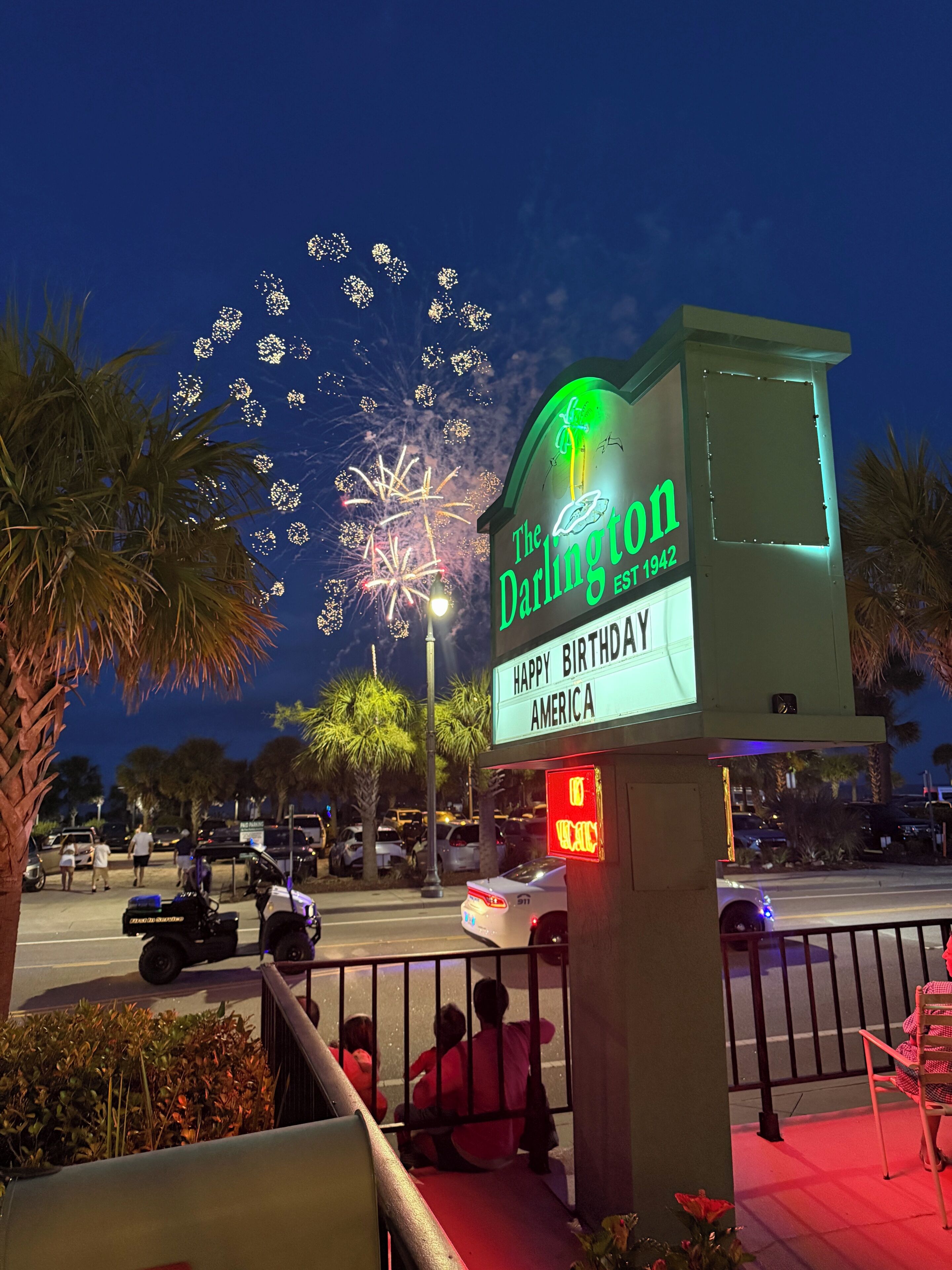 A sign for 'The Darlington' with a 'Happy Birthday America' message, palm trees, and a street view with parked cars and a police SUV.