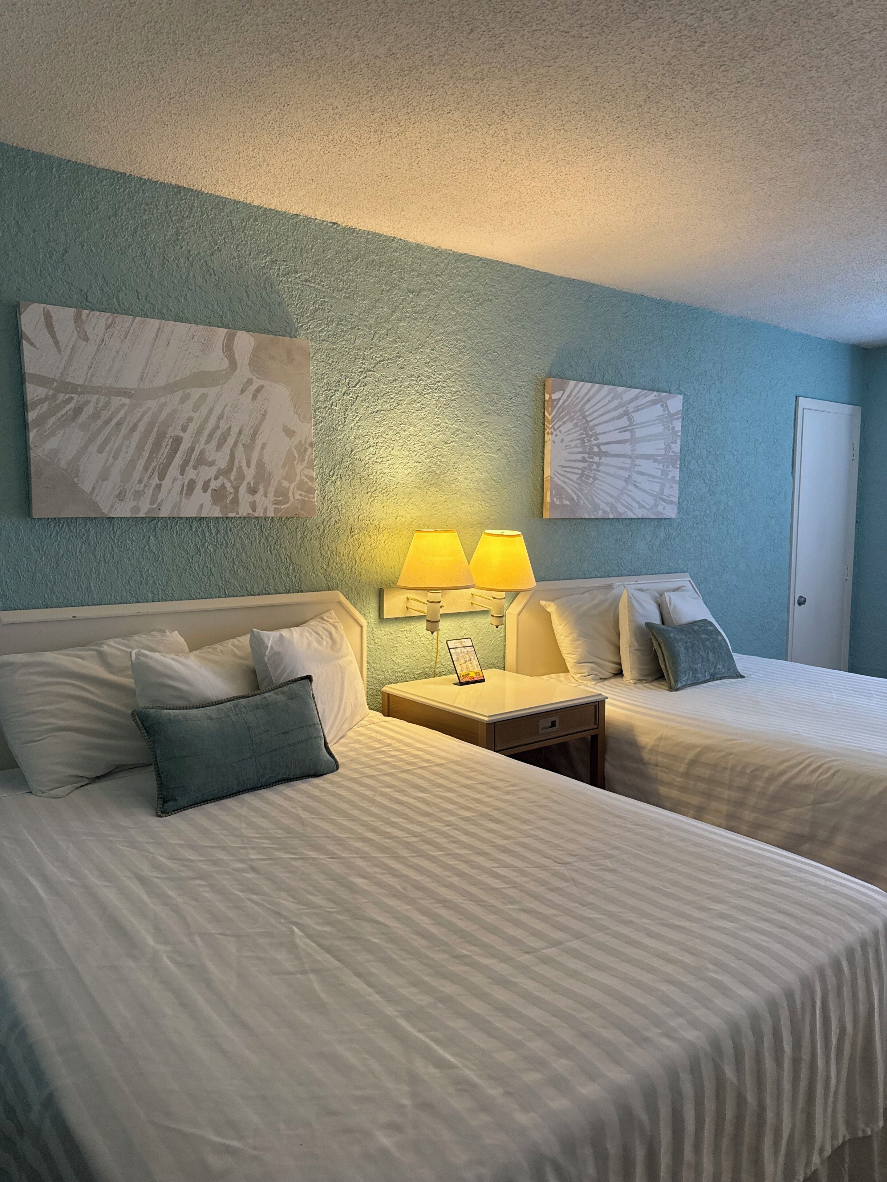 A hotel room with two beds, a nightstand with lamps, and artwork on the walls.