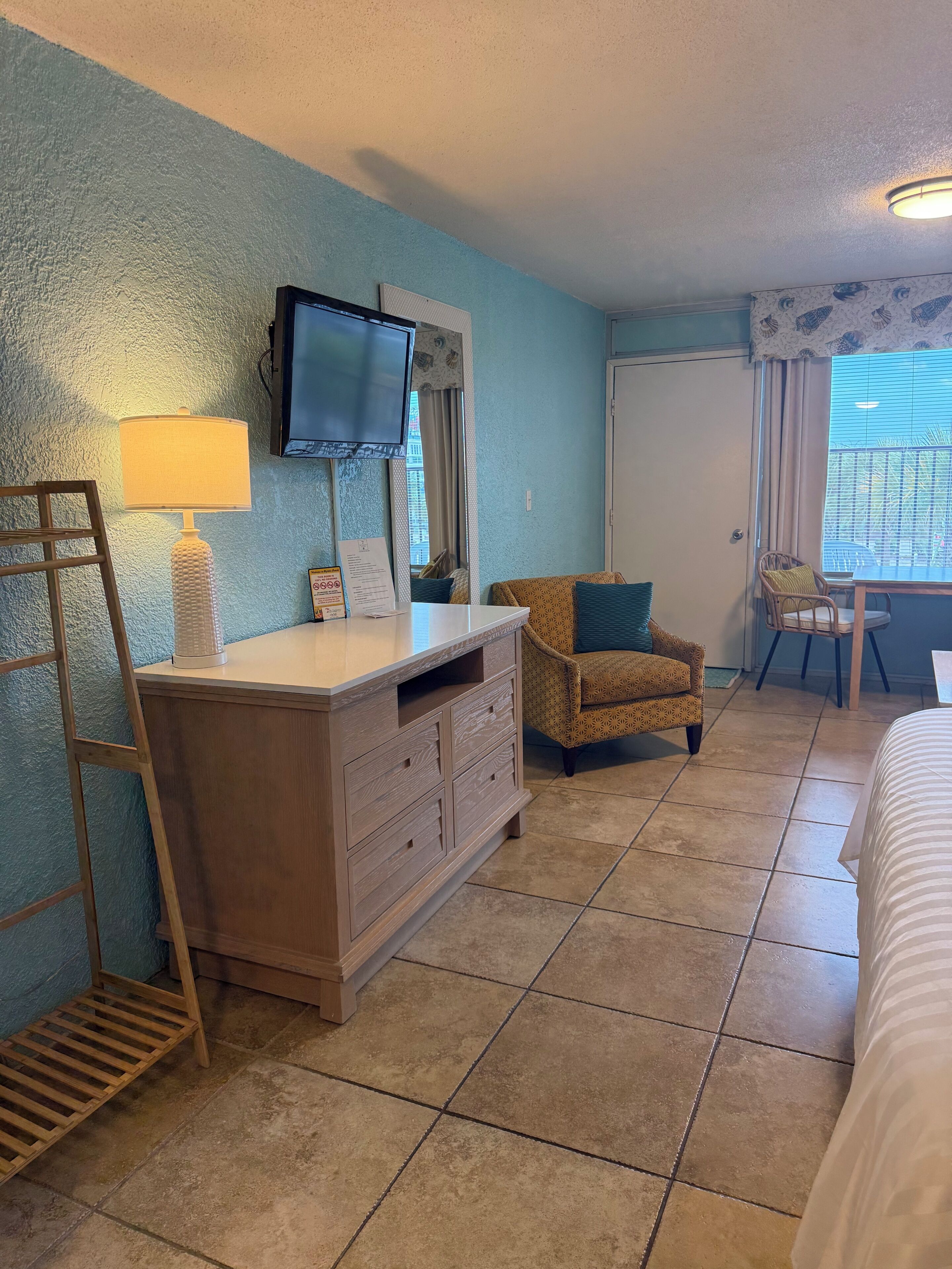 A room with a wooden dresser, a television mounted on the wall, a chair, a bed, and a window with blinds.