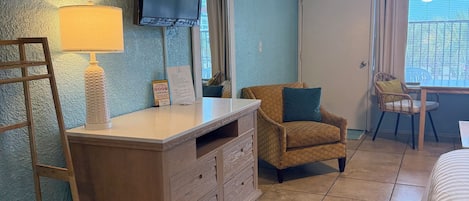 A room with a wooden dresser, a television mounted on the wall, a chair, a bed, and a window with blinds.