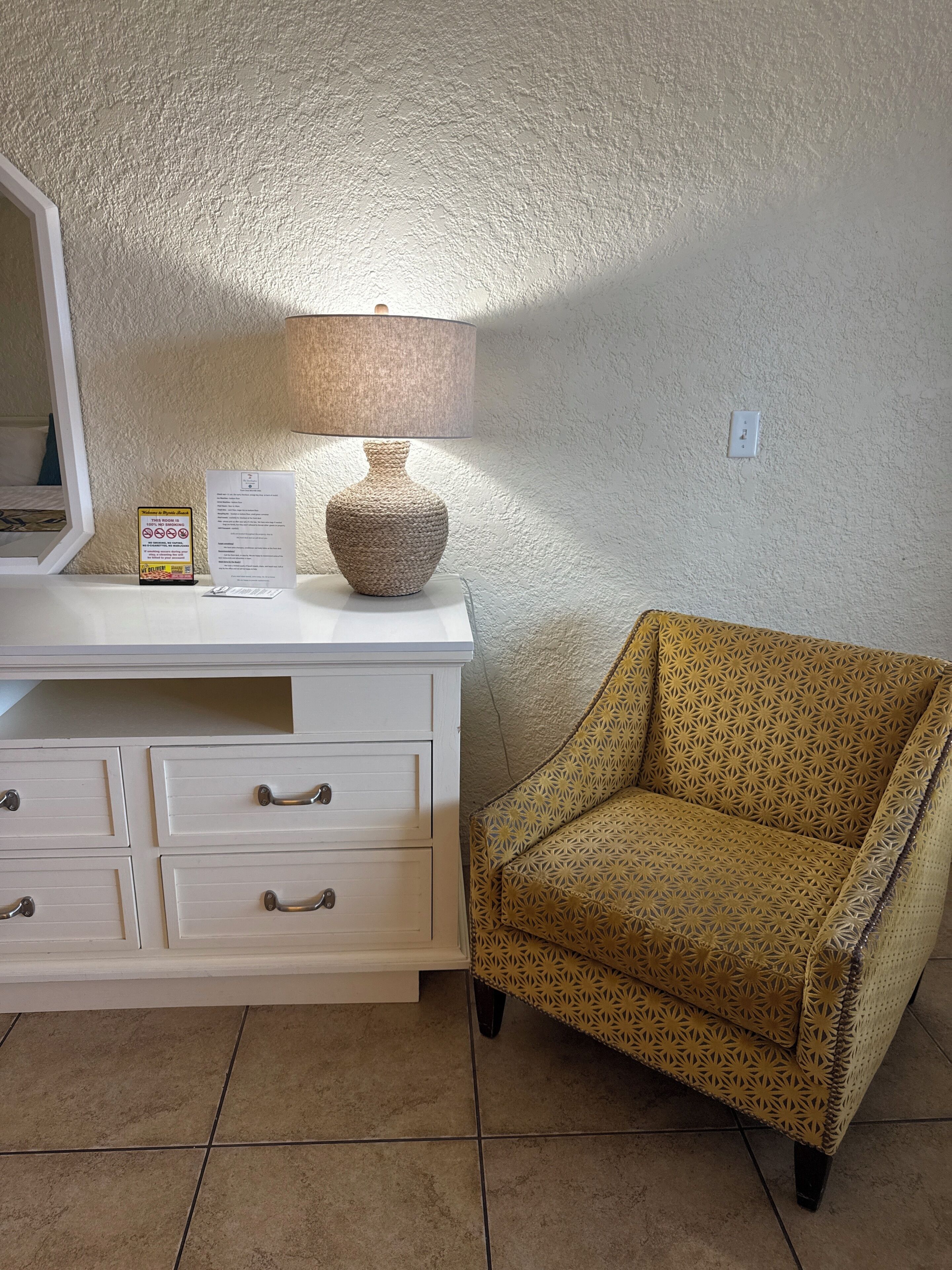 A room with a white dresser, a patterned armchair, a lamp, and a sign on the dresser.