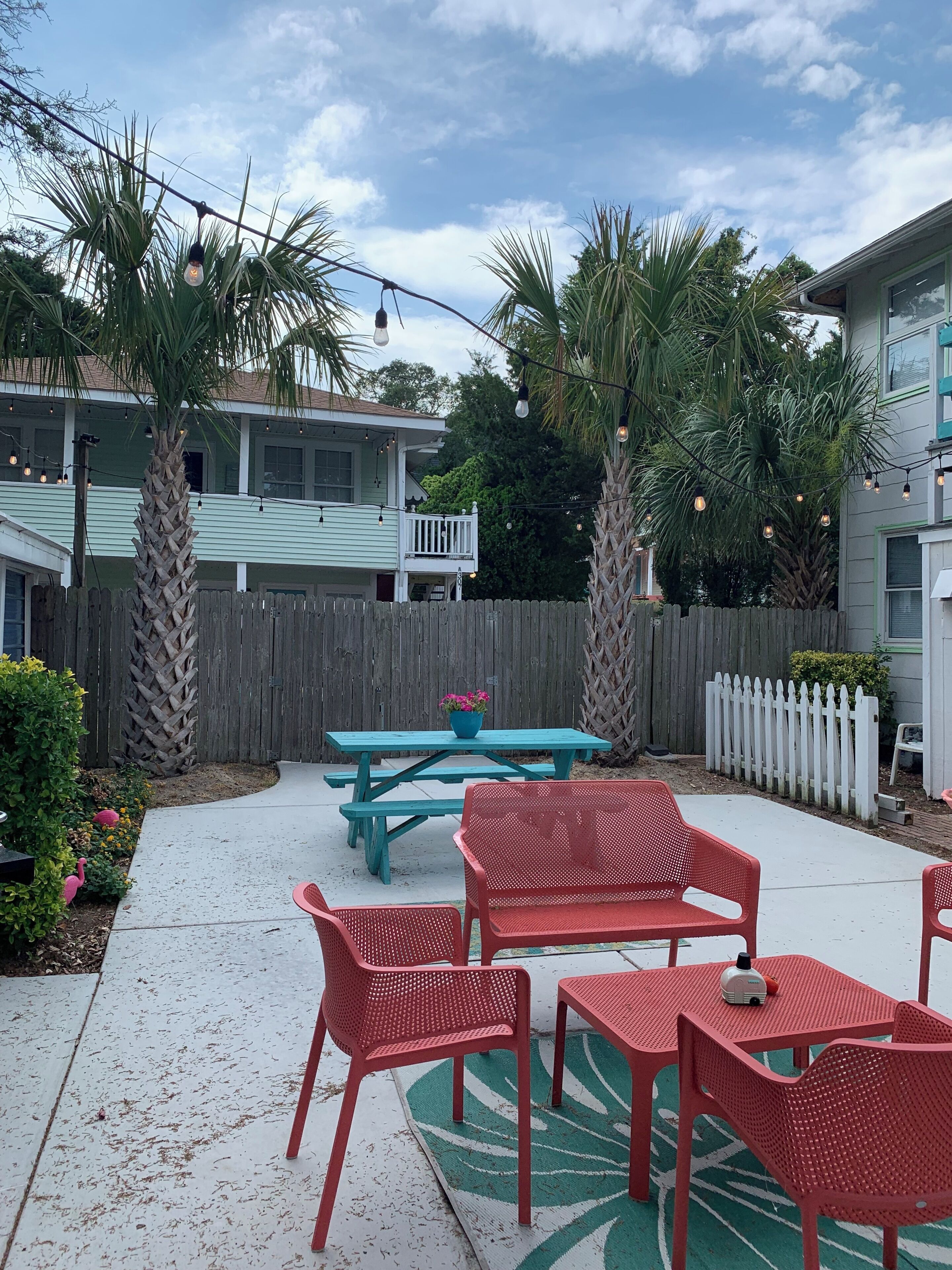 A patio with a picnic table and red chairs, surrounded by a wooden fence and palm trees.