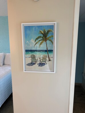 A framed painting of a beach scene with a palm tree and two chairs. - Darlington Inn (Myrtle Beach)