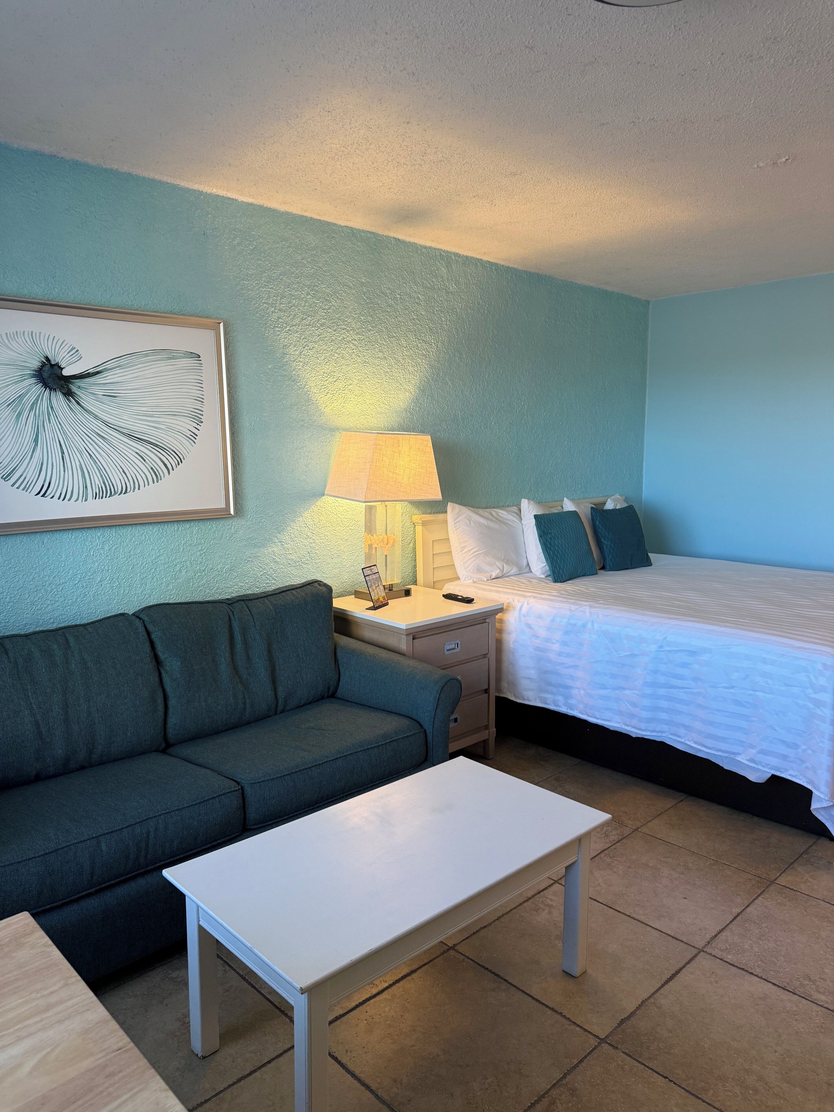A room with a bed, a sofa, a nightstand, a lamp, and a framed artwork on the wall.