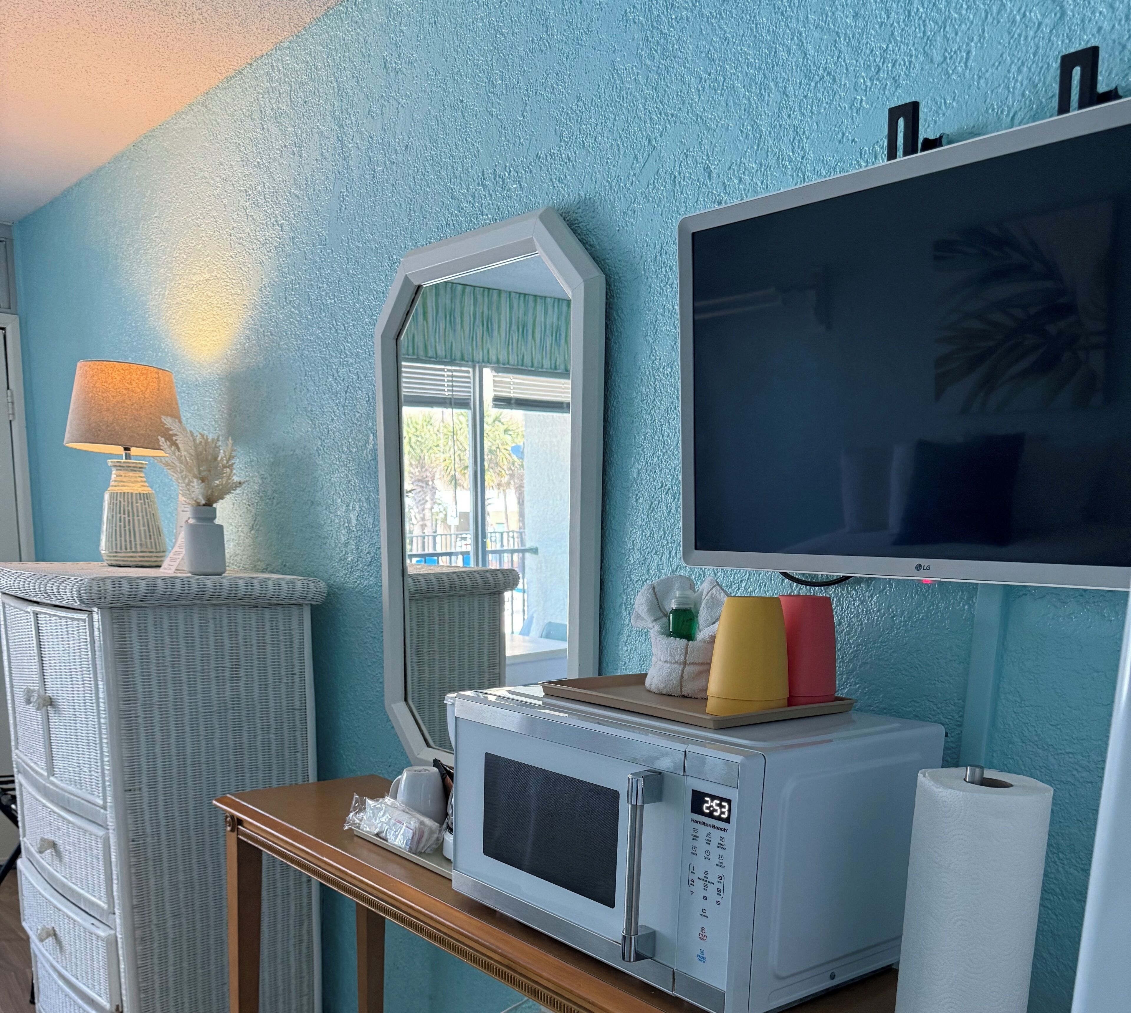 Una habitación con pared azul con textura, un armario de mimbre, microondas, una mesa con lámpara, espejo y televisor.