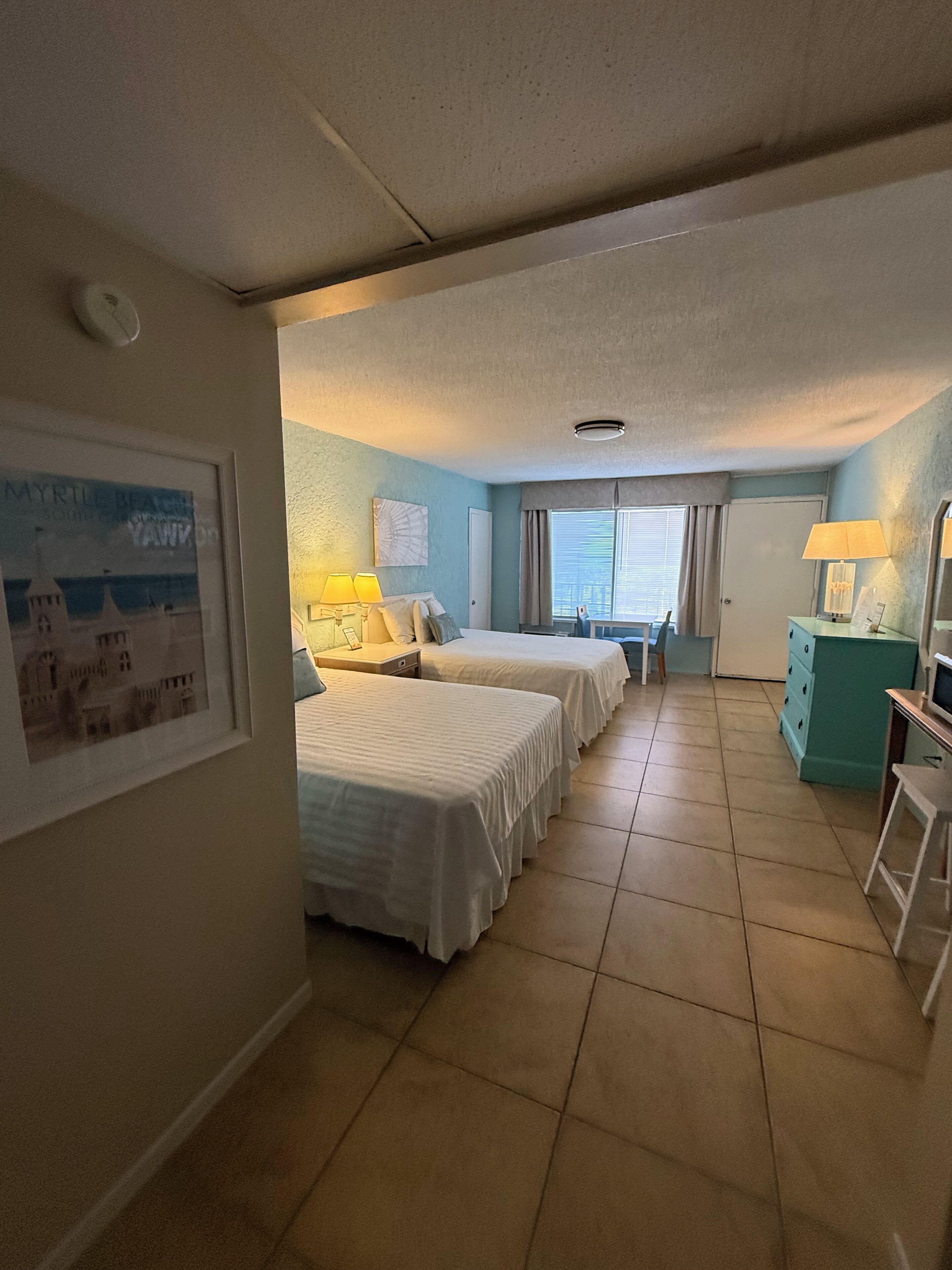Habitación de hotel con dos camas, un escritorio, una silla, un aparador y una ventana con cortinas.