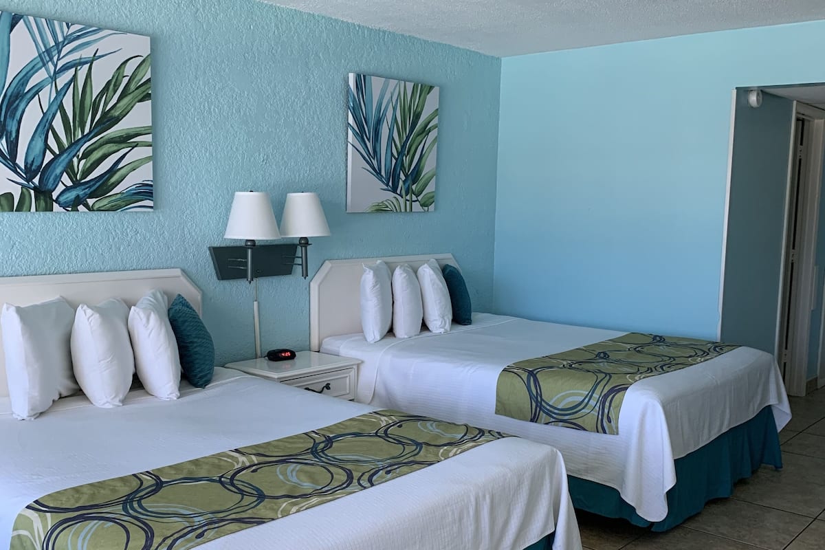 A hotel room with two beds, each with a white bedspread and teal skirted. There are two wall-mounted lamps and two large plant prints on the walls.