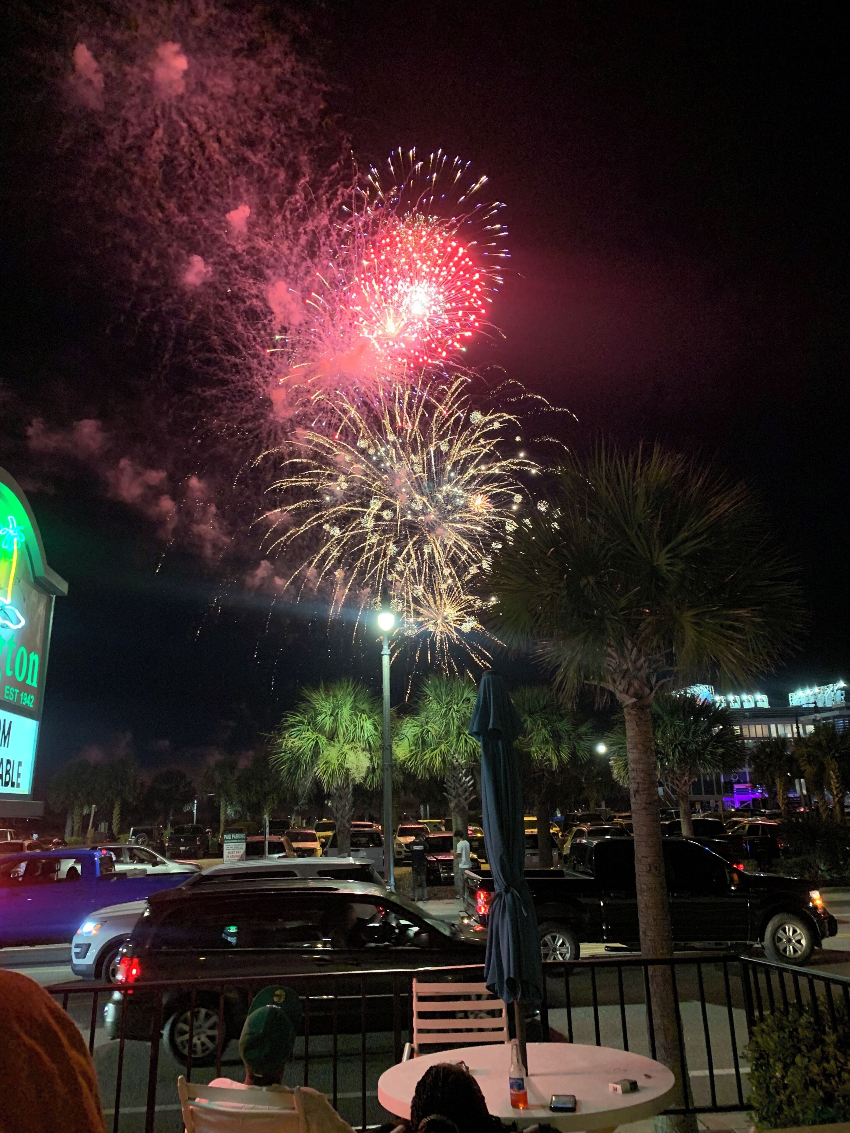 A nighttime scene with fireworks, a parking area, and a building with a neon sign.